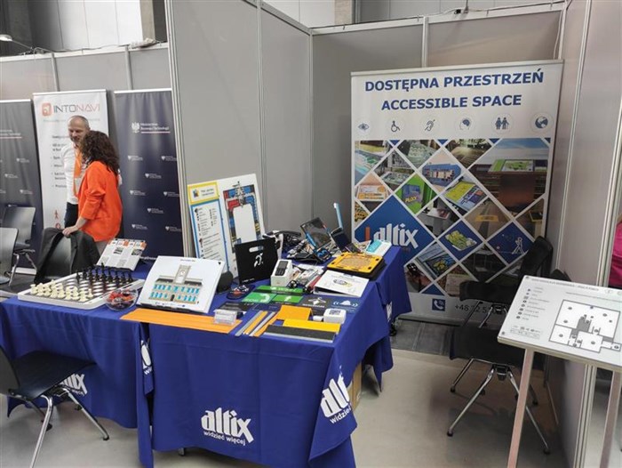 Stoisko wspólne firmy Altix oraz Ministerstwa Rozwoju i Technologii. Po lewej stronie para pracowników Ministerstwa na pierwszym planie Mówiące Szachy, oznaczenia poziome, plany tyflograficzne oraz kilka innych produktów z oferty firmy Altix. w W tle rollup z napisem: Dostępna Przestrzeń Accessible Space.