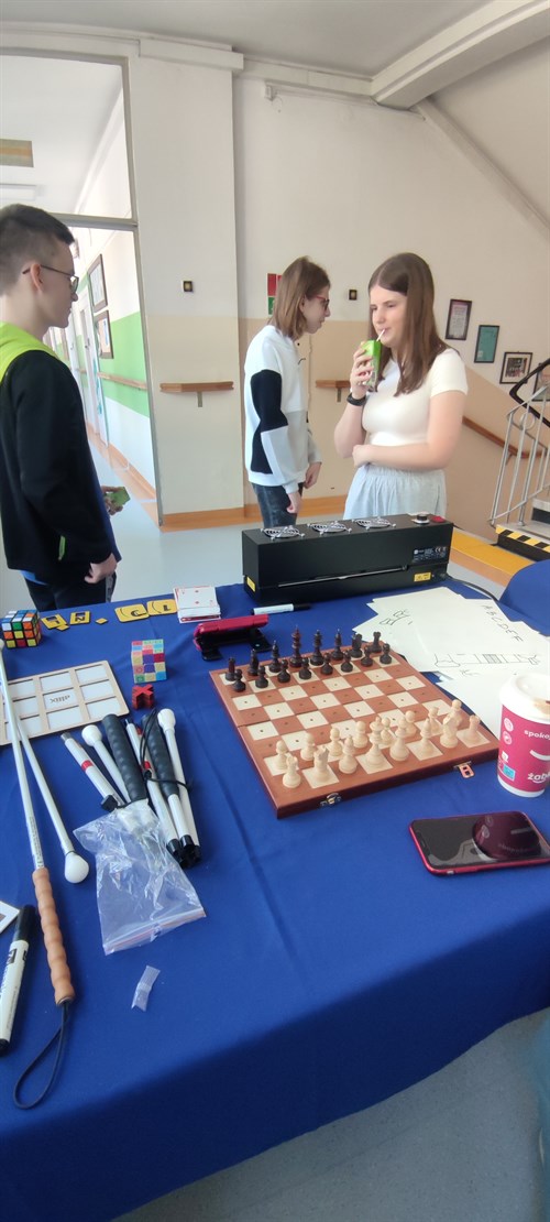 Na pierwszym planie znajduje się stół z niebieskim obrusem, na którym znajdują się sprzęty z oferty firmy Altix takie jak białe laski, drewniane gry (szachy, kółko i krzyżyk) oraz wygrzewarka Zy-Fuse. Na drugim planie widać trójkę uczniów szkoły.