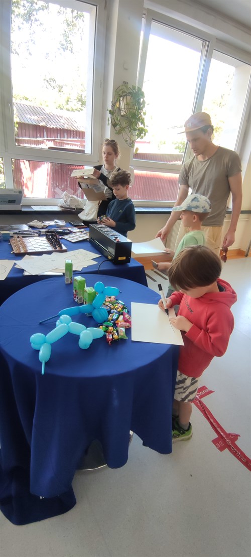 Na pierwszym planie przy okrągłym stoliku z niebieskim obrusem mały chłopiec rysuje. Na stoliku znajdują się baloniki w kształcie pieska oraz słodycze. W tle przy drugim stole znajduje się dwójka dzieci oraz para dorosłych, którzy pomagają dzieciom wygrzać ich rysunki na papierze pęczniejącym.