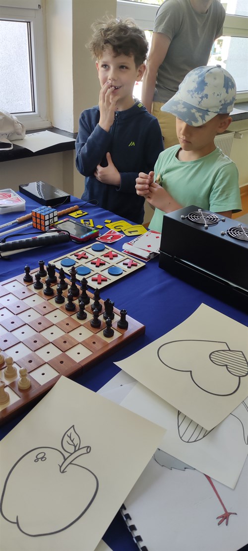 Na pierwszym planie znajdują się przygotowane wcześniej na papierze pęczniejącym ilustracje przedstawiające jabłko, małe serduszko wpisane częściowo w większe serce, a także bociana. Obok rysunków znajdują się szachy, gra drewniana kółko i krzyżyk, kostka Rubika, karty do gry oraz wygrzewarka Zy Fuse. w tle widać dwóch uśmiechniętych chłopców.