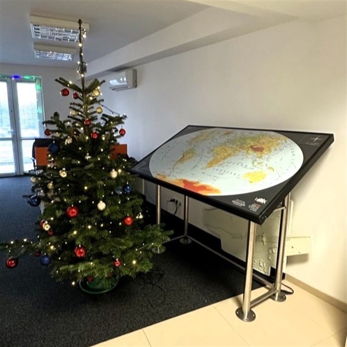 Choinka z bombkami w kolorach czerwony, złoty i granatowy oraz lampkami w kolorze żółtym stoi obok tyflograficznej mapy świata.