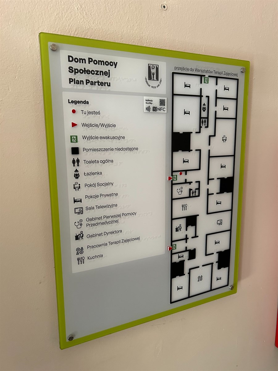 Tablica z planem budynku z wypukłymi symbolami i systemem YourWay Beacon, umieszczona na ścianie przy podłodze z prowadnicami dotykowymi.