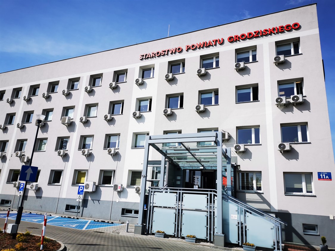 Wejście Główne Do Budynku Starostwa Powiatu Grodziskiego Z Pochylnią I Oznaczonymi Miejscami Parkingowymi Dla Osób Z Niepełnosprawnościami