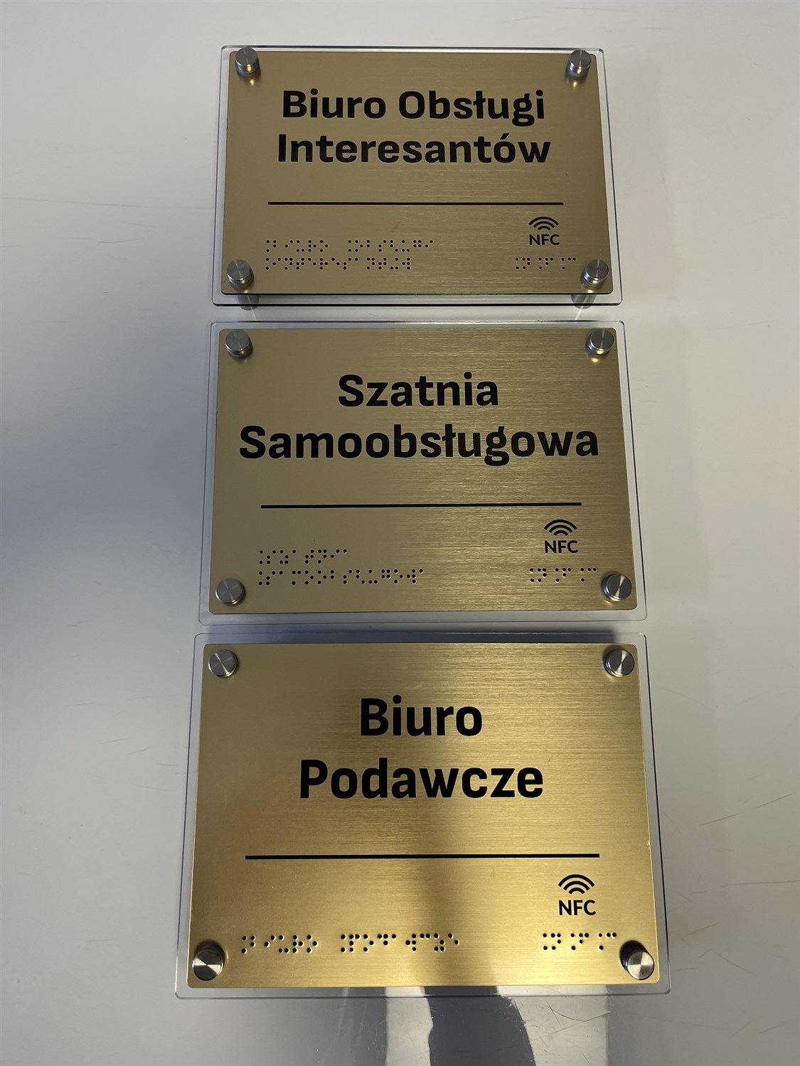 Trzy tabliczki informacyjne z Braille’em i NFC: „Biuro Obsługi Interesantów”, „Szatnia Samoobsługowa” i „Biuro Podawcze”.