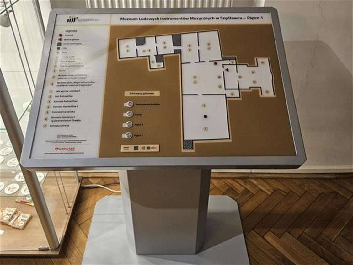 Duży, szary, wolnostojący kiosk multimedialny z wbudowanym udźwiękowionym planem, przedstawiającym rozkład piętra 1 w Muzeum Instrumentów Ludowych w Szybowcu. Plan jest w kolorze brązowym z jaśniejszymi, wypukłymi konturami pomieszczeń. Na tablicy widnieje legenda z symbolami i opisami w Braille'u, a także logo NFC i YourWay Beacon. W tle widać fragment kaloryfera i jasną ścianę.